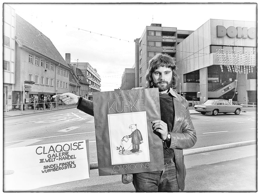 Joachim Kupke mit seinem ersten Cartoon-Band zeigt auf die Claqoise-Boutique, in der nicht nur er, sondern beispielsweise auch Roland Emmerich oder Michel Schilling ihre Kunstwerke präsentieren. Im Hintergrund steht das Domo für eine neue Einkaufs- und Wohnwelt. Bild: Helmut Bail/Archiv P. Bausch