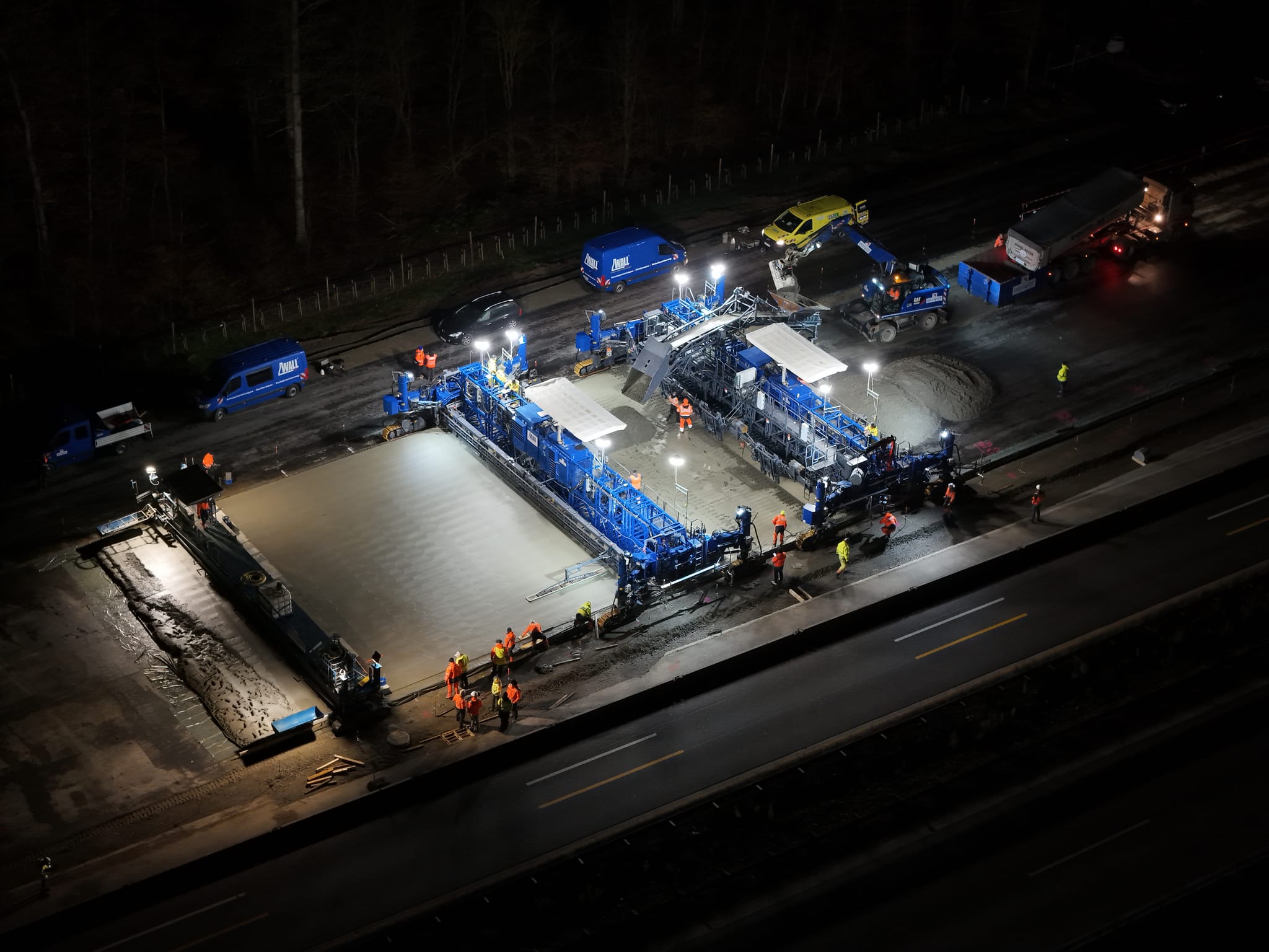 Ein Betonfertiger im Einsatz, der sich von Osten nach Westen vorarbeitet. Der Beton wird in zwei Lagen aufgebracht, verteilt, verdichtet und geglättet.