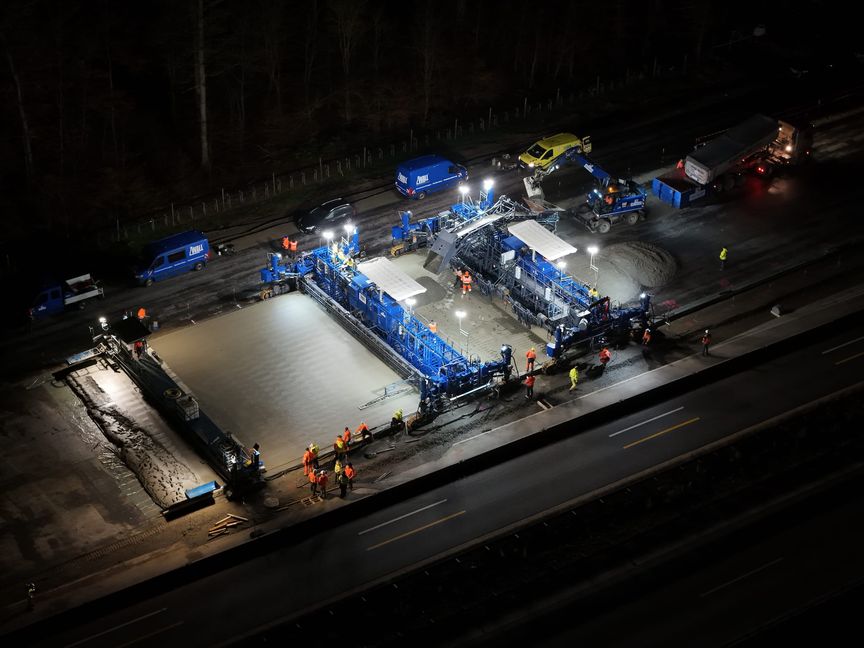 Ein Betonfertiger im Einsatz, der sich von Osten nach Westen vorarbeitet. Der Beton wird in zwei Lagen aufgebracht, verteilt, verdichtet und geglättet.