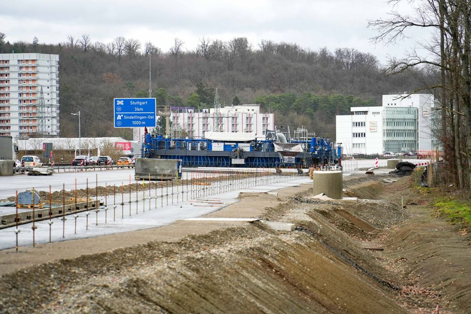 Die dunkle Fläche ist die Asphalttragschicht, auf die die Betondecke mithilfe des Betonfertigers aufgetragen wird. Der Betonfertiger arbeitet sich von Osten nach Westen vor.