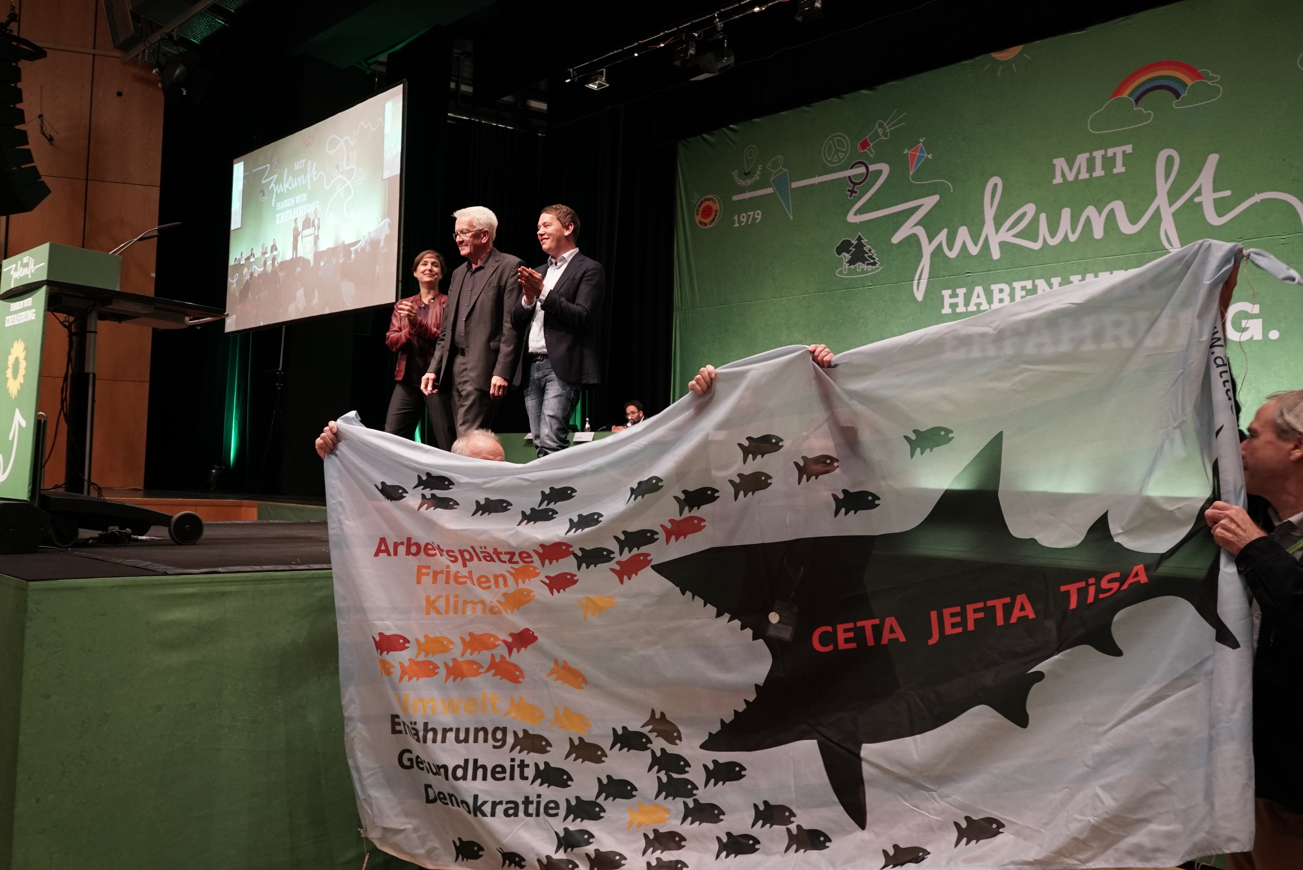 Winfried Kretschmann nimmt gerahmt von Sandra Detzer und Oliver Hildenbrand Applaus für seine Rede entgegen, während Aktivisten mit Plakaten gegen das CETA-Freihandelsabkommen demonstrieren. Bild: Heiden