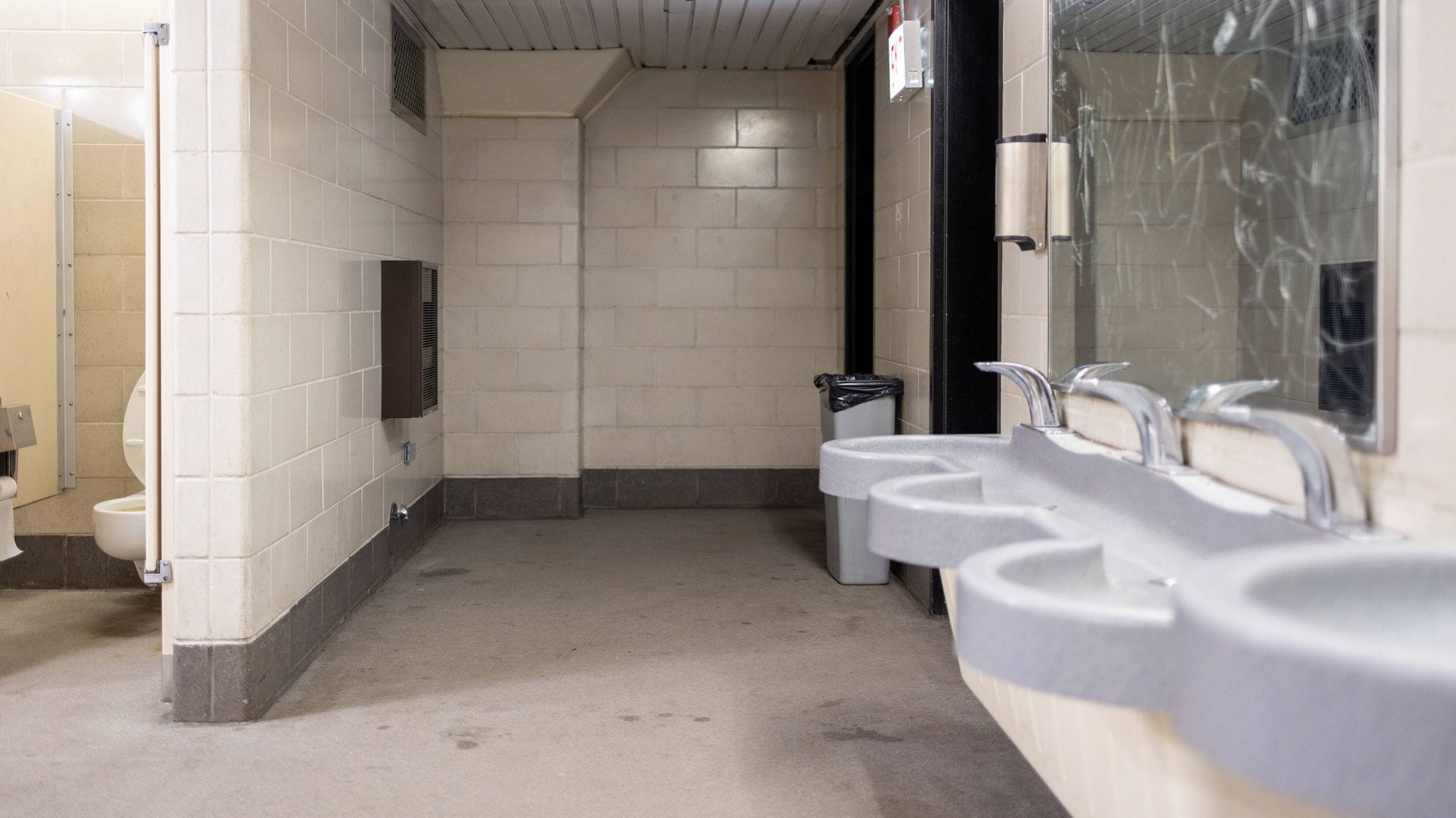 Innenansicht einer öffentlichen Toilettenanlage mit Waschbecken, Urinalen und einzelnen WC-Kabinen. Der Raum ist funktional gestaltet mit Fliesenwänden, Trennwänden und standardisierten Sanitäranlagen. Mulmig ist es einem trotzdem, wenn man an einem solchen Ort muss.