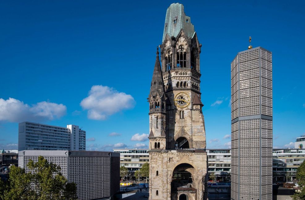 Die Kaiser-Wilhelm-Gedächtnis-Kirche gehört zu den bekanntesten Wahrzeichen Berlins (Foto vom 11.10.2021). Die Einweihung der alten Kirche im neoromanischen Stil fand am 1. September 1895 statt. Im Zweiten Weltkrieg wurde die Kirche durch einen Bombenangriff im Jahr 1943 stark beschädigt. Nur die Turmruine blieb danach als Mahnmal gegen den Krieg erhalten. Zwischen 1959 und 1963 wurde der alte Turm durch ein vierteiliges Gebäudeensemble des Architekten Egon Eiermann  ergänzt. Dazu zählen das am 17. Dezember 1961 eingeweihte achteckige Kirchenschiff, der sechseckige Glockenturm, die viereckige Kapelle und das Foyer.
