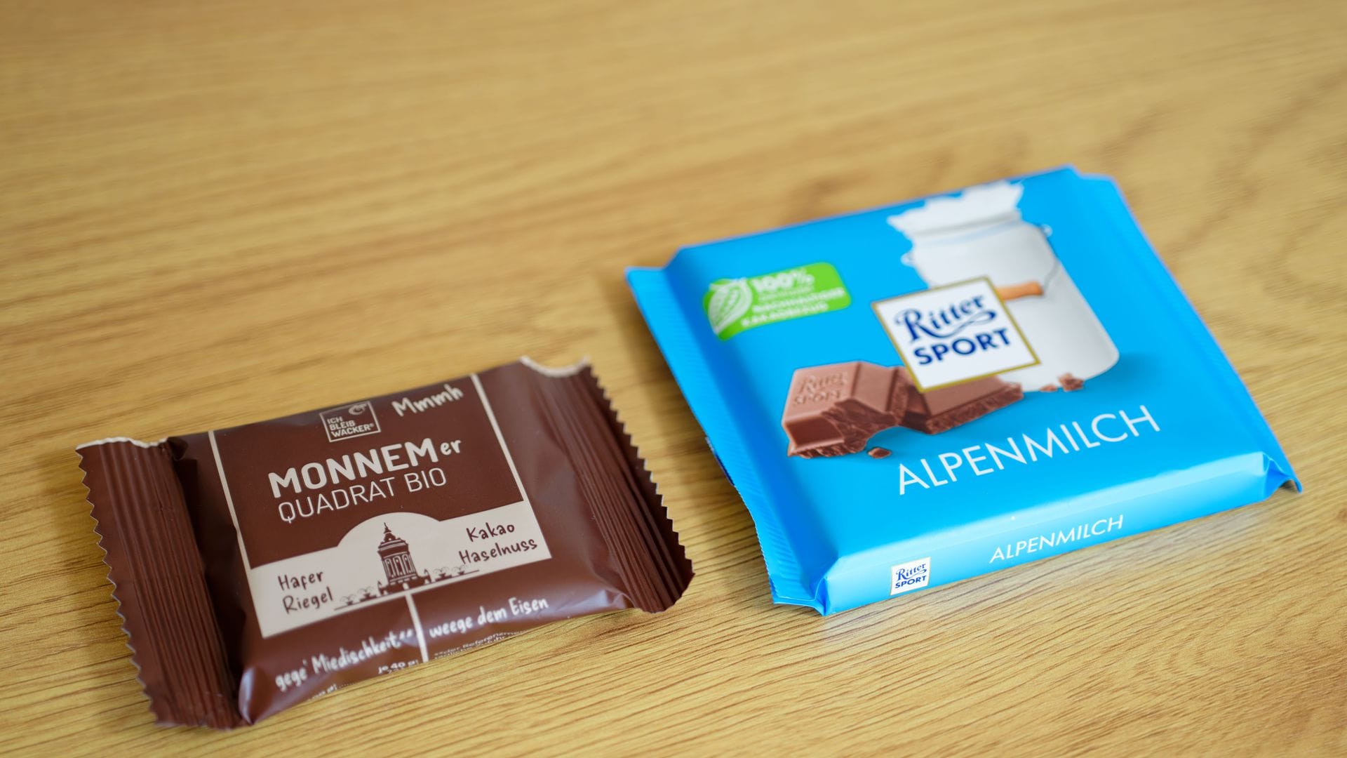 Eine Tafel mit Schokolade der Marke Ritter Sport (r) und ein Haferriegel der Firma Wacker mit der Aufschrift „Monnemer Quadrat Bio“ liegen nebeneinander auf einem Tisch.