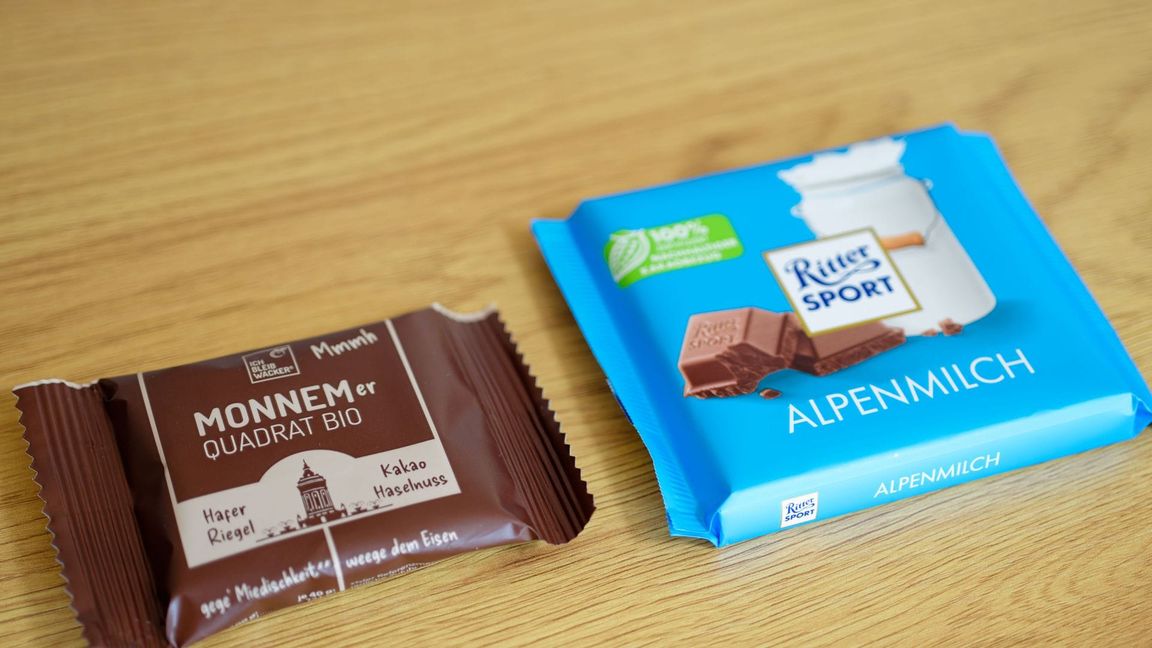 Eine Tafel mit Schokolade der Marke Ritter Sport (r) und ein Haferriegel der Firma Wacker mit der Aufschrift „Monnemer Quadrat Bio“ liegen nebeneinander auf einem Tisch.