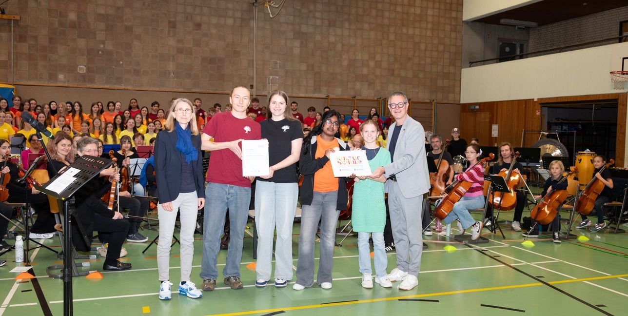 Schulleiterin Nadine Kußler (links) und Kulturamtsleiter Markus Nau mit den Schülern Ruben Walker, Lilly Kasnar, Sruthi Prathaban, Bernice Deubler bei der Überreichung der Plakette im Rahmen einer Gesamtprobe für das Musical „One sun, one world“.