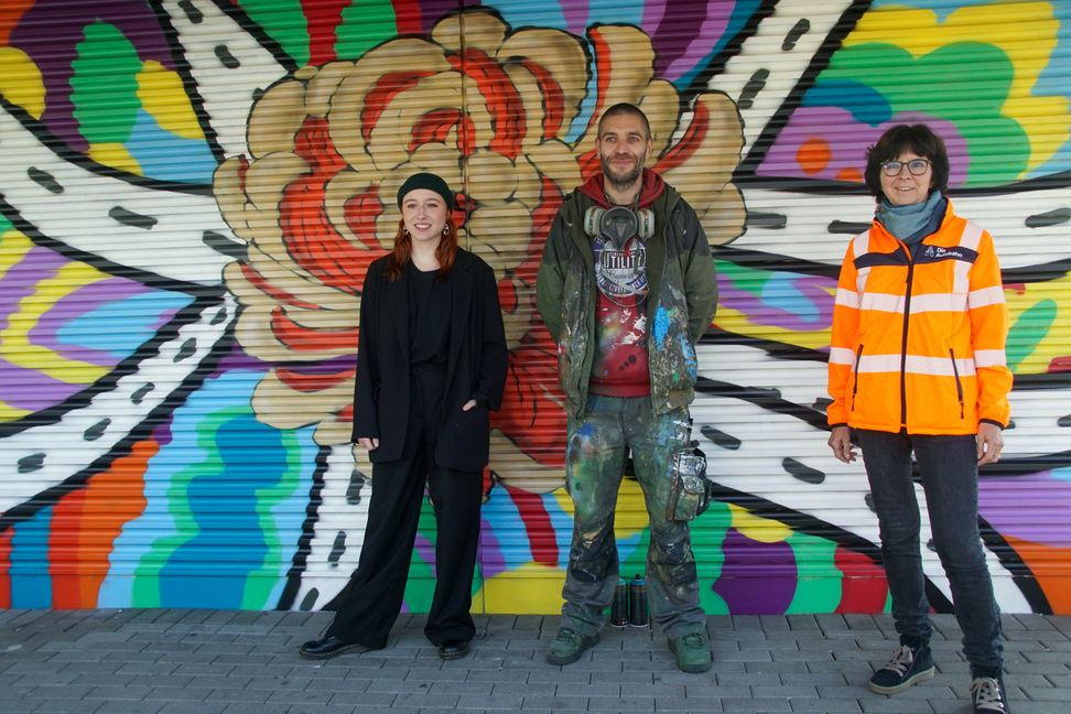 Gewinnerin Johanna Munke, Sprayer Marco Billmaier und Niederlassungsdirektorin Christine Baur-Fewson (von links) vor der neu besprühten Wand.