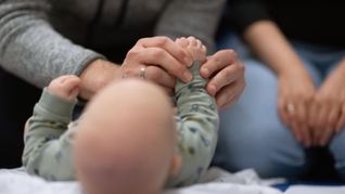 Ein Vater hält während eines Infant Handling Kurses die Füße seines Kindes, während die Mutter daneben sitzt.