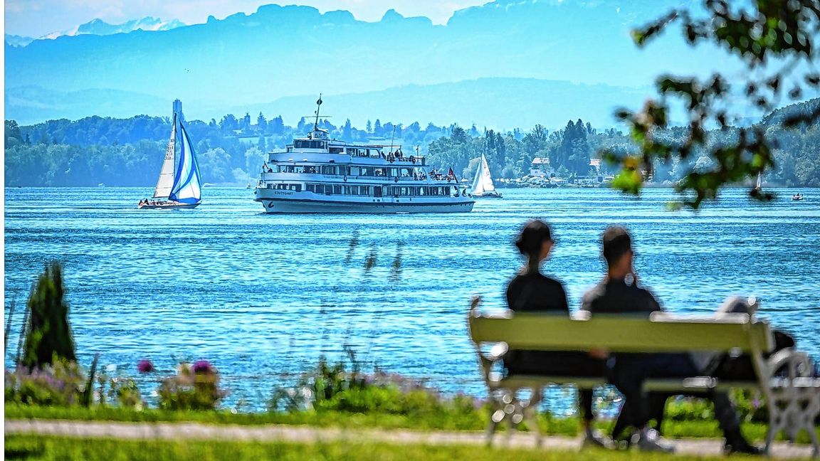 Das Passagierschiff Stuttgart fährt auf dem Bodensee zur Anlegestelle Überlingen, während im Vordergrund ein Paar auf einer Bank im Uferpark sitzt.