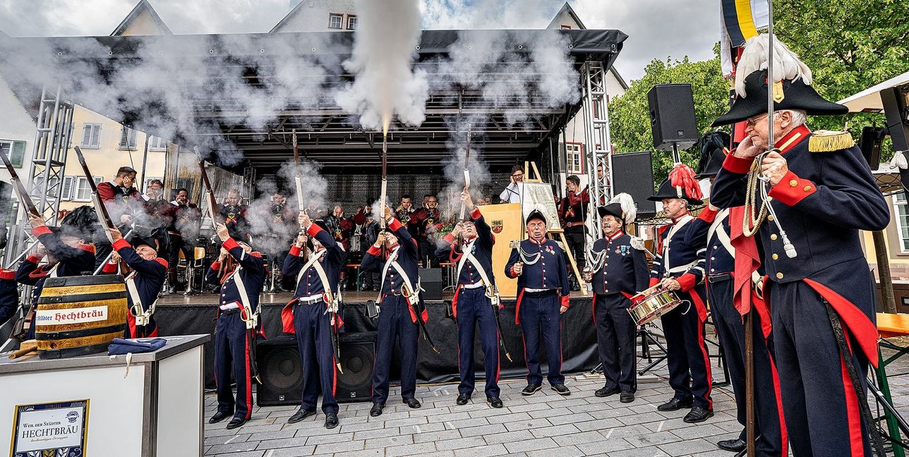 Drei Schuss aus dem Gewehr als Salut zum Stadtjubiläum in Weil der Stadt. Rechts Bürgerwehr-Kommandeur Hartmut Sigel. Bürgermeister Christian Walter am Rednerpult hält sich die Ohren zu. Dahinter der Fanfarenzug.