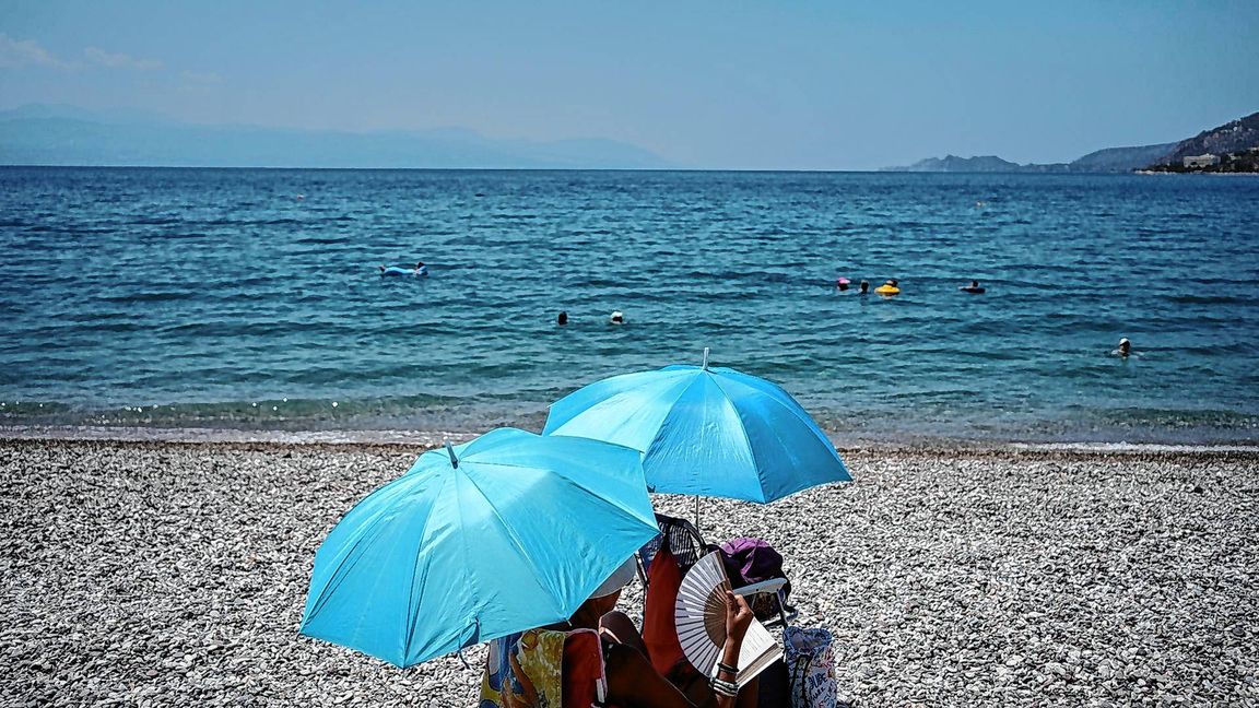 Eine Frau mit einem Handventilator unter einem Sonnenschirm am Strand.