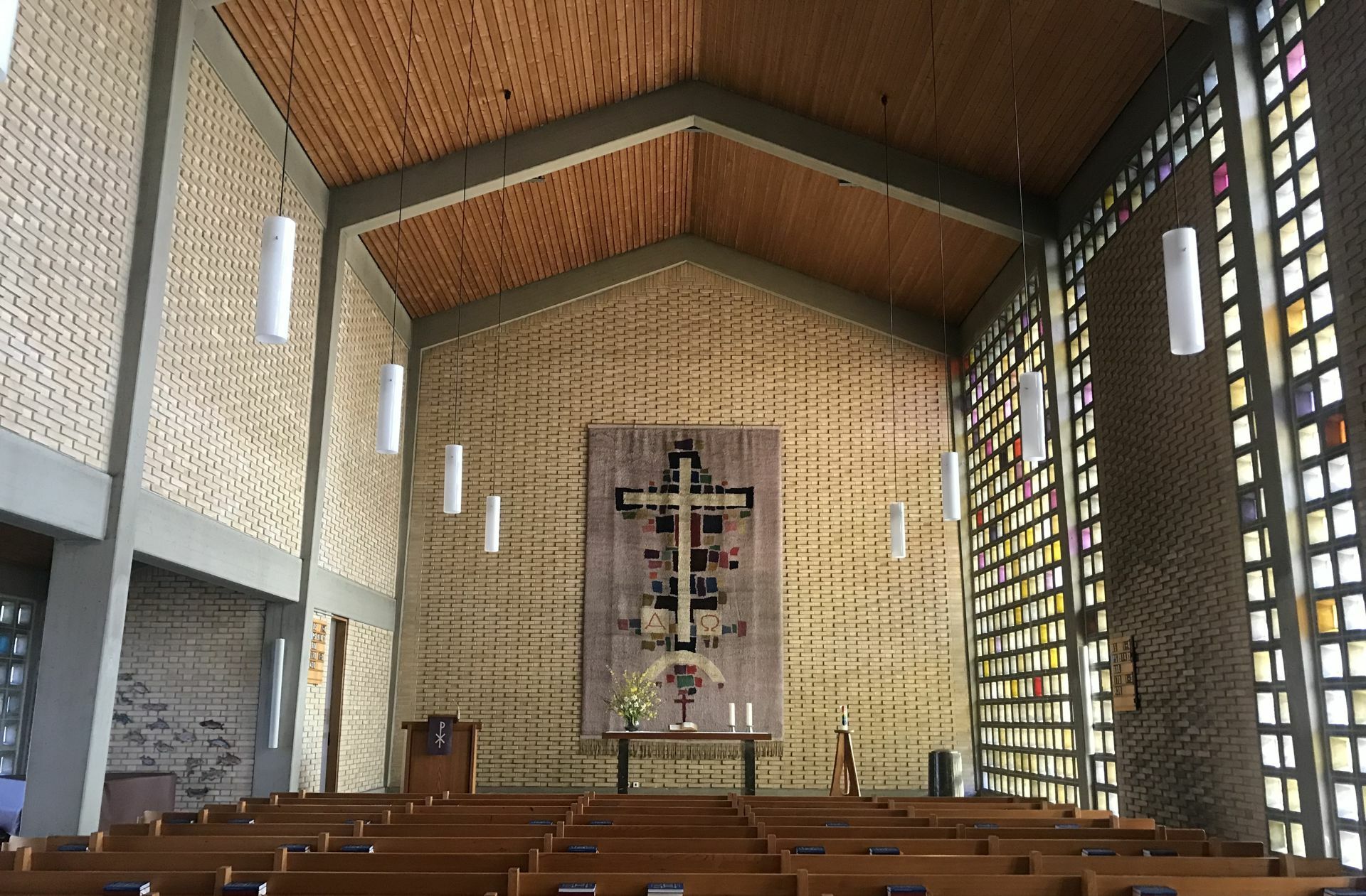 Der Innenraum der Kreuzkirche hat eine beeindruckende Atmosphäre. Vor allem, wenn die Sonne durch die Seitenwände des Kirchenschiffes fällt. Sie bestehen aus vielen kleinen Fenstersteinen, die regelrecht zu glühen scheinen, wenn das Licht hindurch strahlt. Besonders die Südwand, deren Steine in warmen Farben gehalten sind und in die drei große Steinmauern eingelassen sind, sticht hervor.