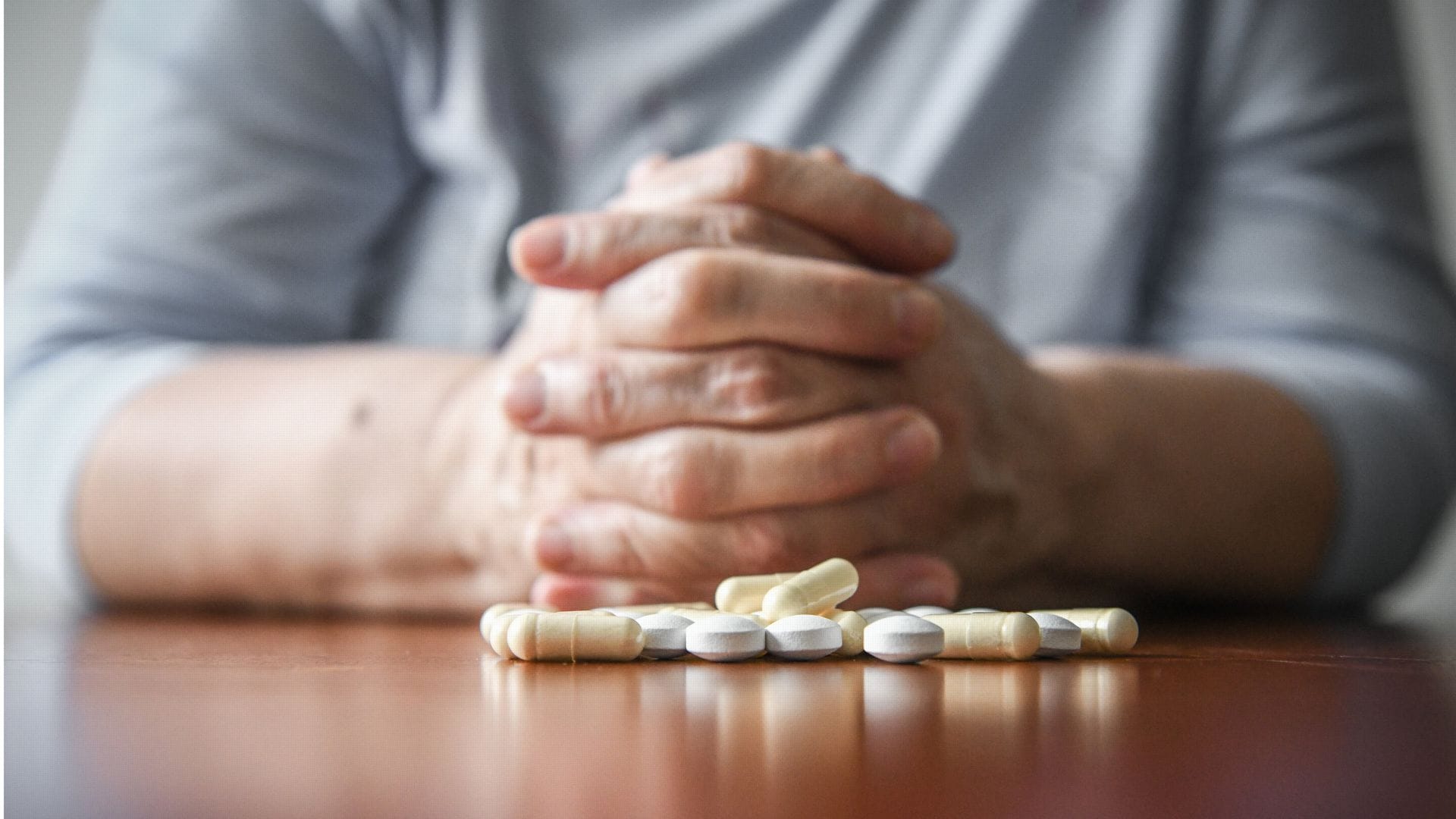 Eine Frau sitzt mit gefalteten Händen an einem Tisch. Vor ihr liegt ein Haufen Tabletten (gestellte Szene).