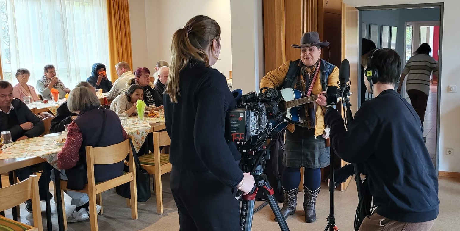 Szene: Jaqueline Bart spielt „Take me Home, Country Roads“ auf ihrer Gitarre, während die Filmcrew sie filmt. Im Hintergrund sitzen die Gäste.Bild: Cardoso
