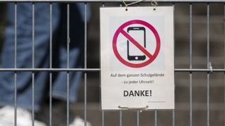 Ein Schild mit einem durchgestrichenen Smartphone und der Aufschrift „Auf dem ganzen Schulgelände - zu jeder Uhrzeit! DANKE!“ hängt am Zugangstor zum Schulhof eines Gymnasiums. (Symbolbild)