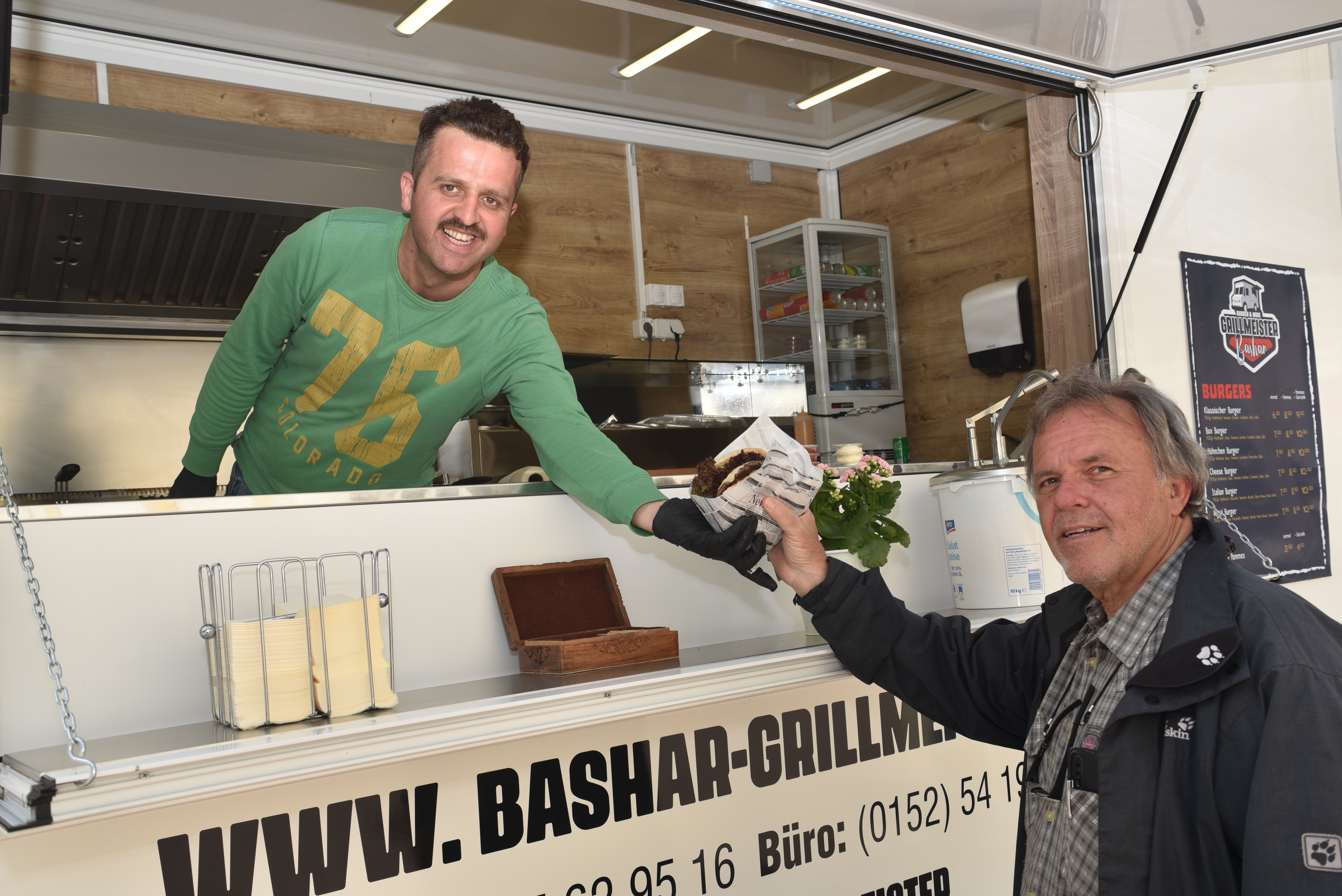 Bashan Kadan (links) in seinem Imbisswagen und sein Förderer Dieter Mast. Den Burger gibt es entweder in Papier oder – zum Mitnehmen – in einem kleinen Karton verpackt.  Bild: Reichert