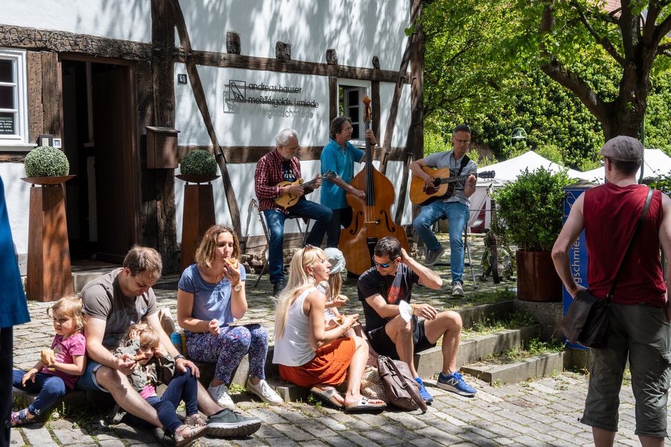 Vor der Goldschmiede von Andreas Hauser spielt er mit seinem Trio „Backporch“, während Besucher des Handwerkermarkts sich einen Snack gönnen und die Musik genießen. Bild: Nüßle