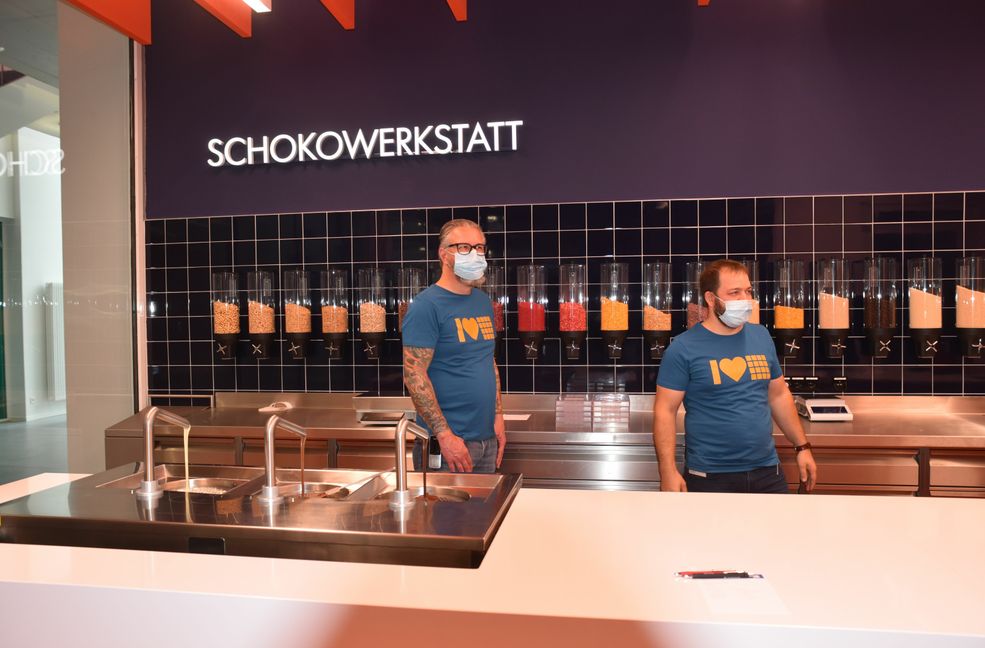 Hier sollen die Mitarbeiter ihre eigene Schokolade kreieren. Die Produktentwickler Joachim Eissner (links) und  Christian Breitbach vor den Zutatenbehältern. Vorne links fließen weiße Schokolade, Milch- und Bitterschokolade aus den Hähnen.               Bild: Reichert